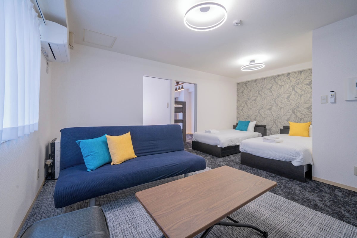 Quarto de hotel com sofá azul, duas camas de solteiro, mesa de madeira e parede estampada.