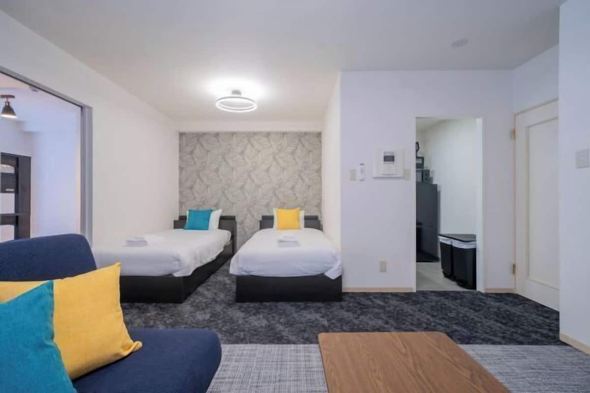 Quarto de hotel com duas camas, um sofá, uma mesa de madeira e um banheiro visível através de uma porta aberta.