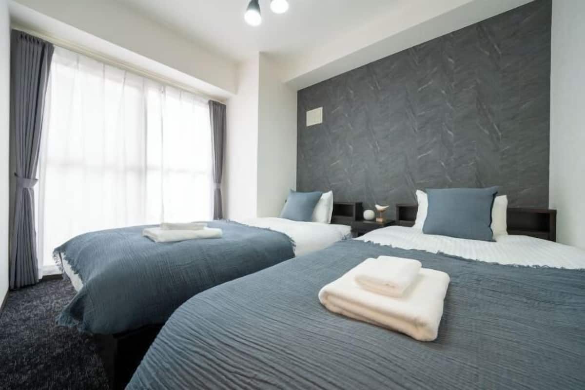 Quarto de hotel com duas camas, roupa de cama azul escura, cabeceira cinzenta e uma janela grande com cortinas.