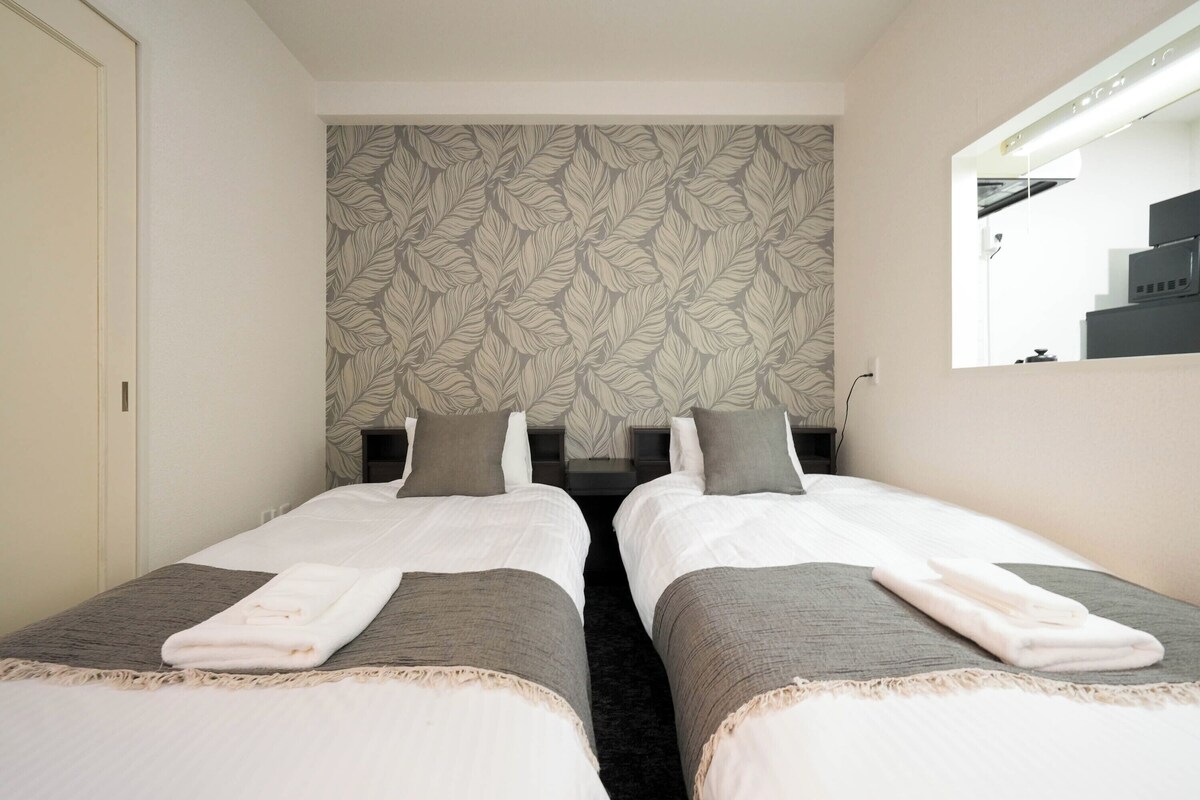 A hotel room with two single beds, a large mirror, a patterned wallpaper, and a view of the kitchen.