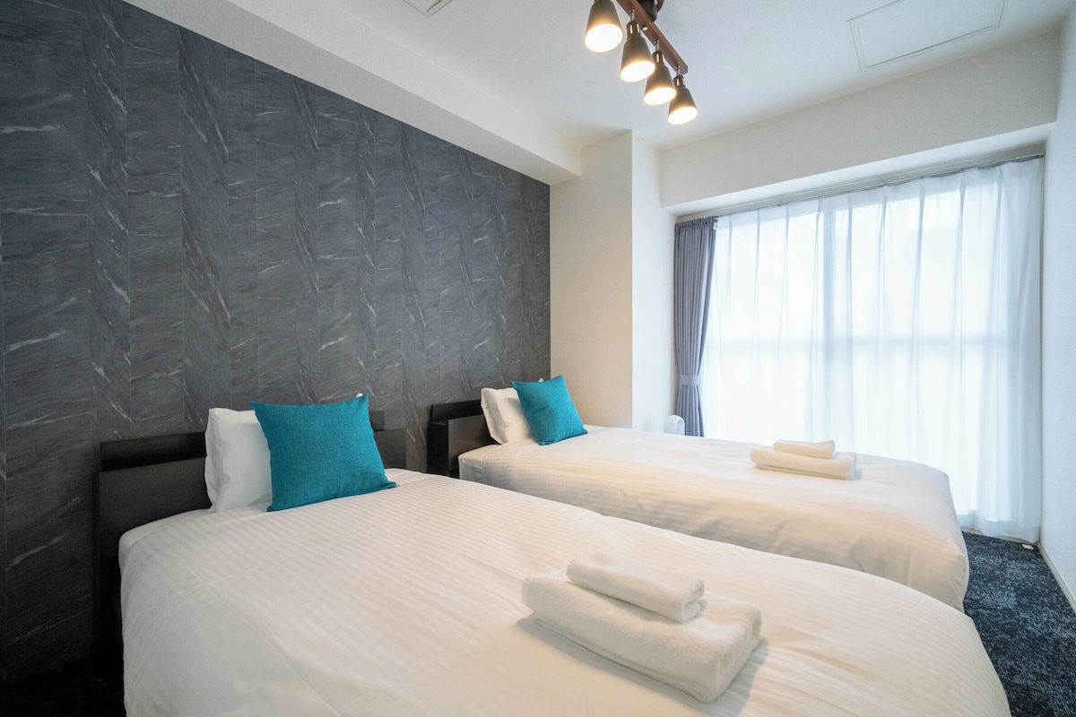 A hotel room with two beds, dark textured walls, a large window with curtains, and a ceiling light fixture.