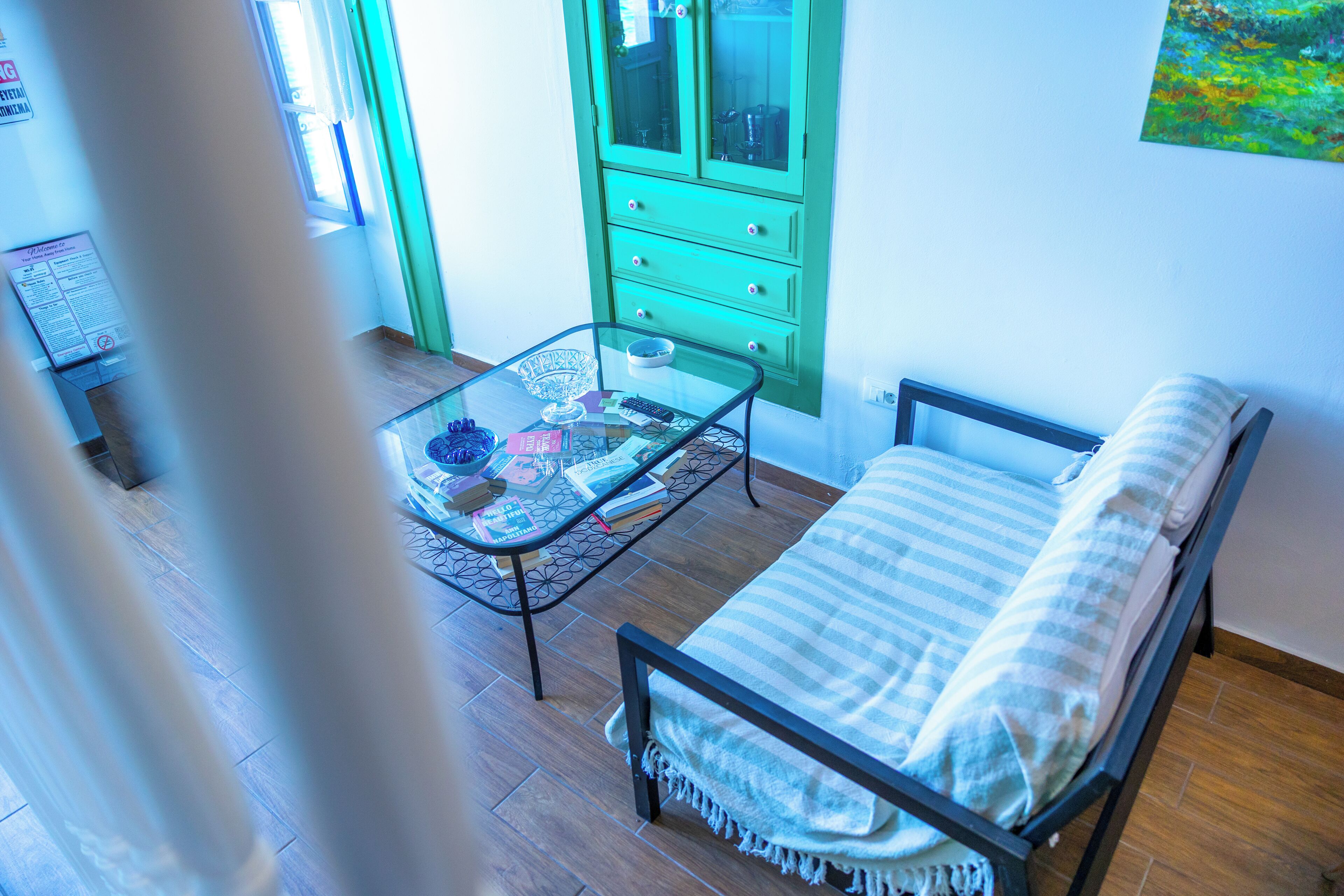 A room with a bed, a glass-top table with books and a bowl, a green cabinet, and a window with curtains.