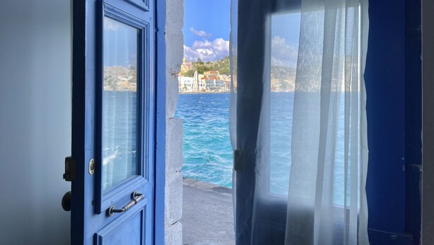 An open door leading to a view of a coastal town with buildings and a body of water. - Pitini 2 - Seafront - Harbour view (Symi)