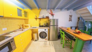 A kitchen with yellow cabinets, a washing machine, a refrigerator, and a dining area with a chair and table. - Pitini 2 - Seafront - Harbour view (Symi)
