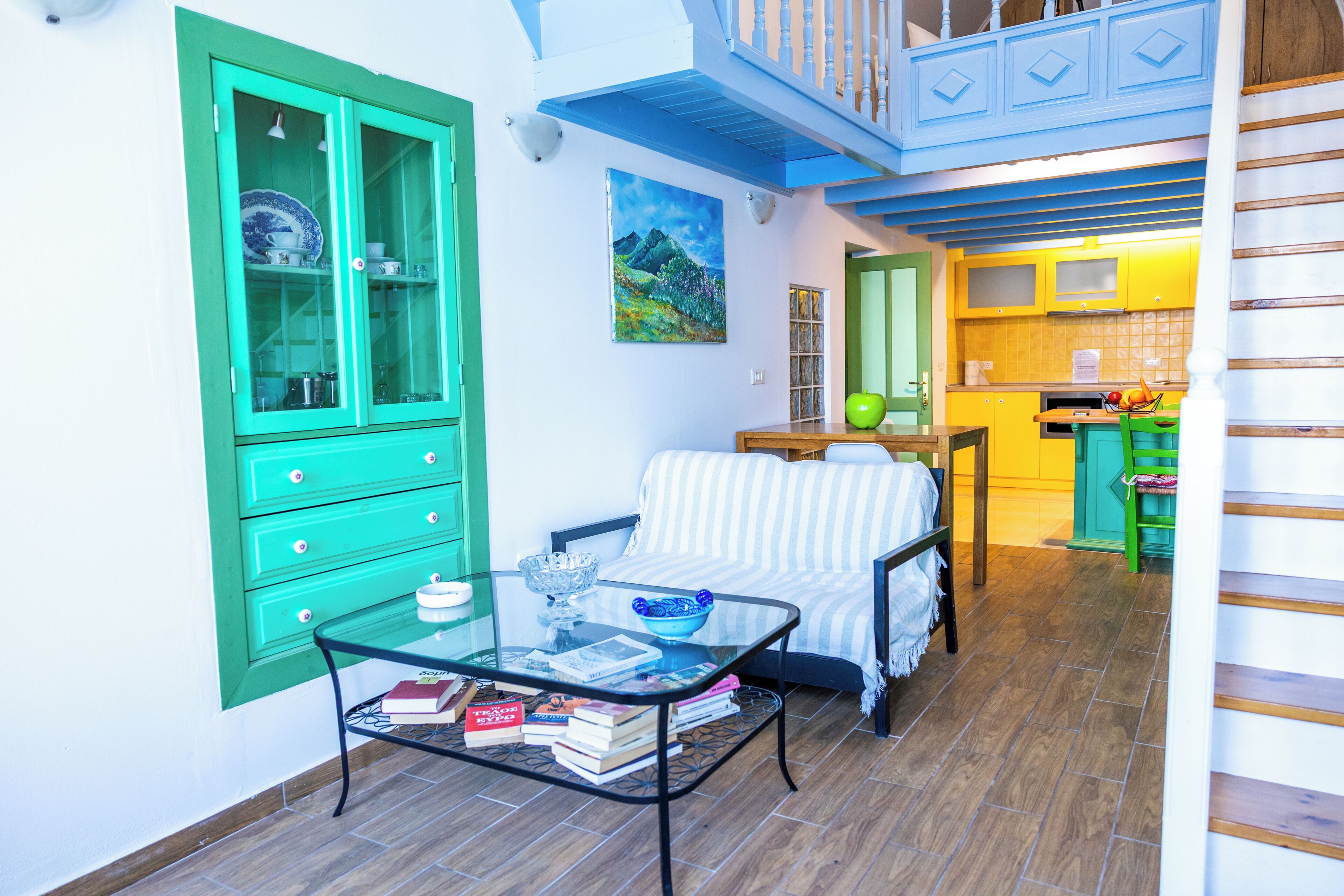 A room with a glass coffee table, a striped sofa, a green cabinet, and a kitchen area with yellow cabinets.