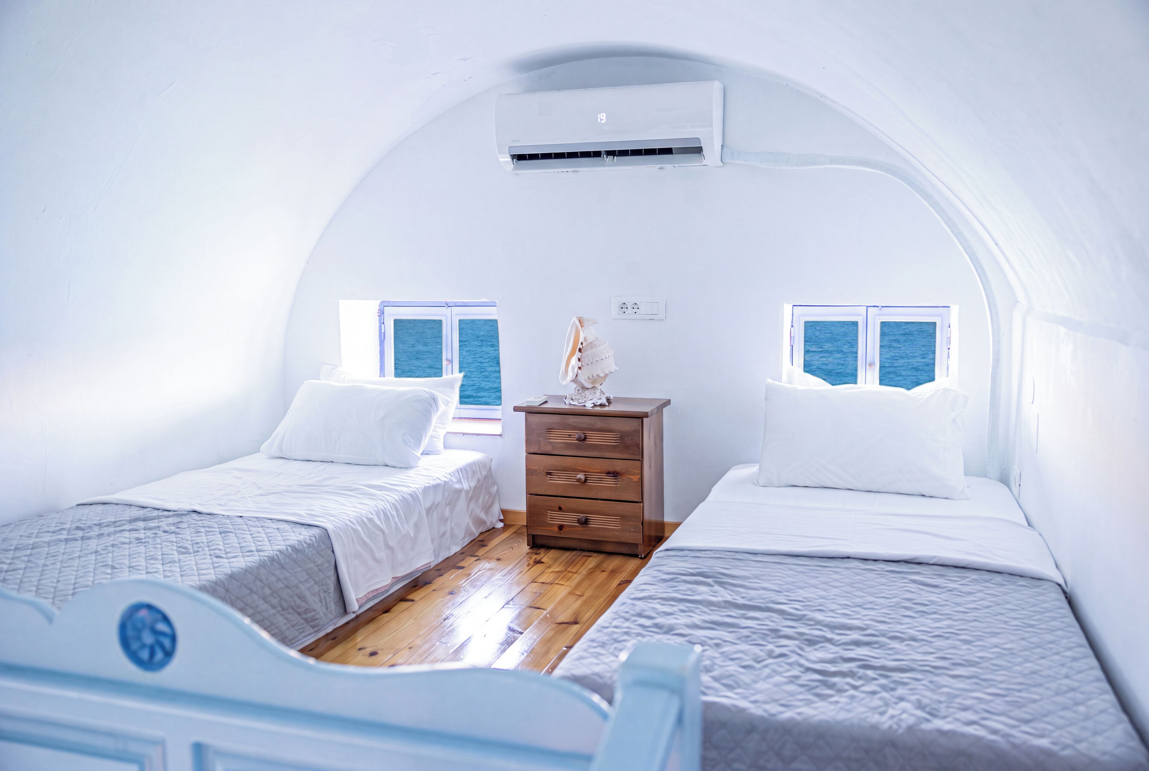 A small, white, dome-shaped room with two beds, a wooden dresser, and a window overlooking the sea.
