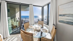 Une pièce avec une table en verre dressée pour le repas, des chaises en osier, une vue sur la plage et un tableau accroché au mur.