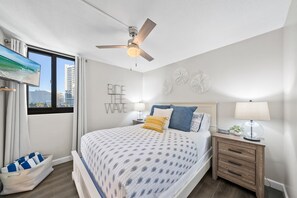 Une chambre à coucher comprenant un lit, un ventilateur de plafond, une table de chevet, une lampe, une fenêtre avec des rideaux et un élément de décoration murale.