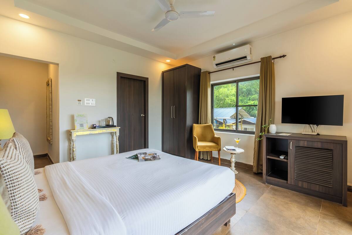 A hotel room with a large bed, a desk with a lamp, a chair, a television, and an air conditioning unit.