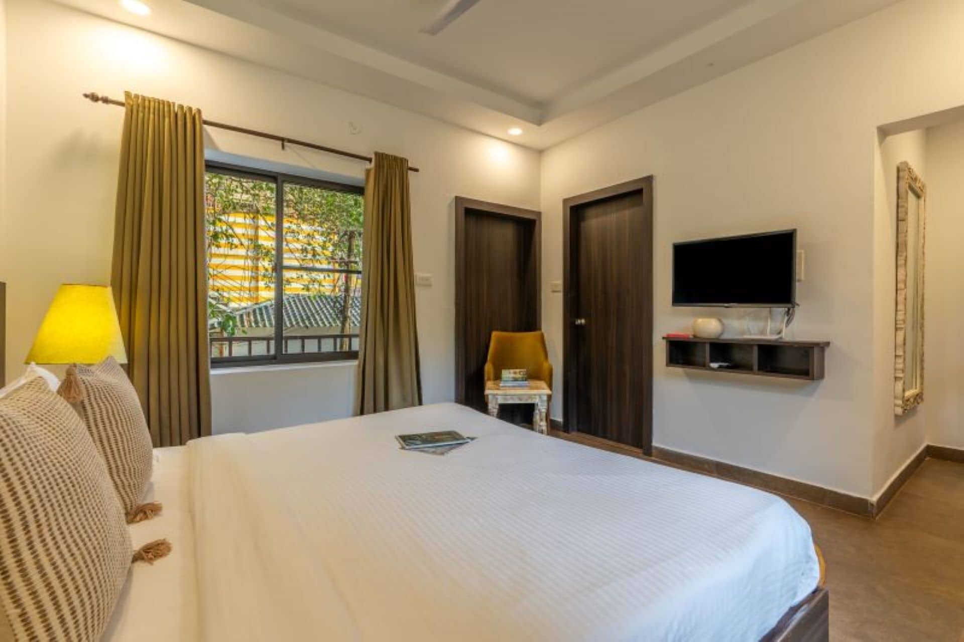 Een hotelkamer met een bed, een aan de muur bevestigde televisie, een raam met gordijnen en een klein tafeltje met een stoel.