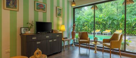 Una sala de estar con televisor, sofá, sillones y vista a una piscina y follaje.