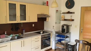 Una cocina compacta con lavamanos, estufa, horno y un refrigerador pequeño. Hay armarios con puertas de vidrio y un reloj en la pared.