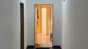 Un pasillo estrecho con suelo de baldosas, una puerta a la izquierda y otra al final que da a un cuarto con piso de madera.