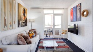 A modern living room with a white sofa, colorful artwork, a flat-screen TV, and a large window.