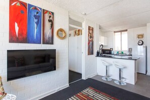 Et moderne køkken med hvide skabe, en sort bordplade og en indbygget mikrobølgeovn. Der er et stort fladskærms-tv monteret på væggen, samt en lille bar med to barstole.