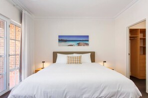 A neatly made bed with white linens and a single decorative pillow, a painting of a coastal landscape above it, and a wooden wardrobe to the right.