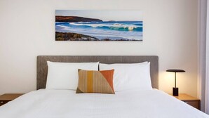 Une chambre d’hôtel avec un lit, une table de chevet, une lampe et un tableau représentant un paysage côtier.