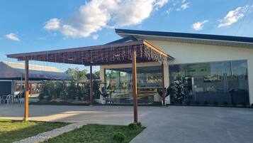 Un área de estar al aire libre con techo, una pérgola, un edificio de cristal con un letrero visible y un césped bien cuidado.