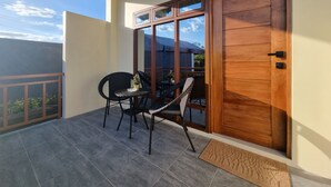 Un balcón con dos sillas y una mesa, una puerta de madera con un cartel encima, y una vista del cielo y las nubes.