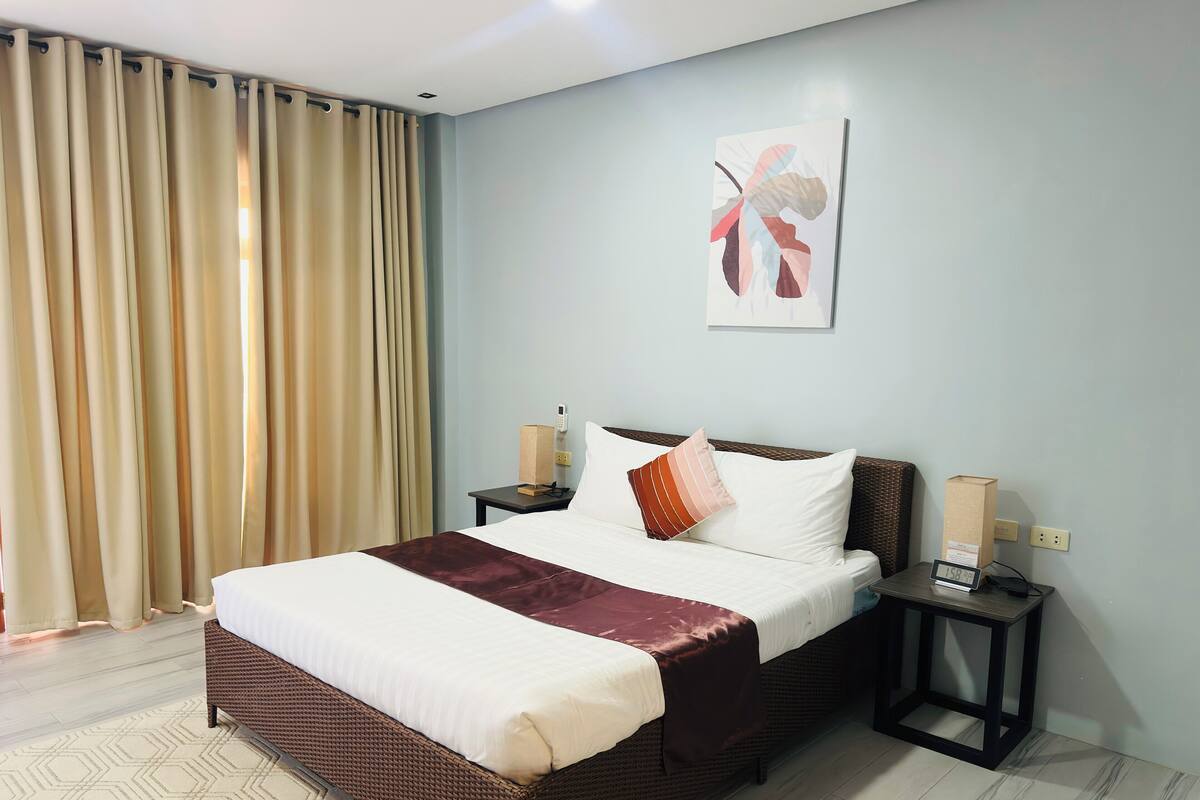 A hotel room with a large bed, a bedside table with a phone and a clock, a wall-mounted artwork, and a window with curtains.