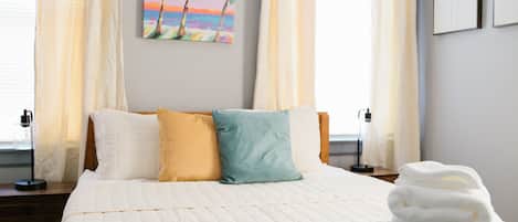 A neatly made bed with a quilted white duvet, two pillows in yellow and blue, and a stack of white towels on the bed.