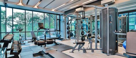 A modern gym with various exercise equipment, large windows, and a bench area.