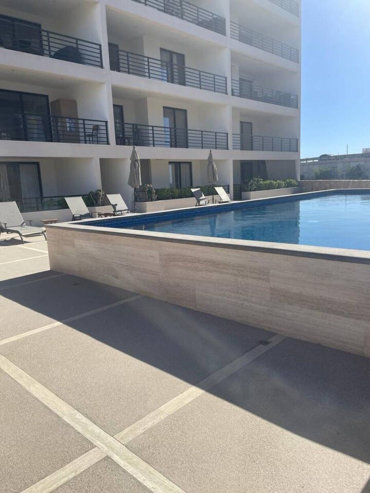 A swimming pool with lounge chairs and umbrellas, surrounded by a multi-story building with balconies.