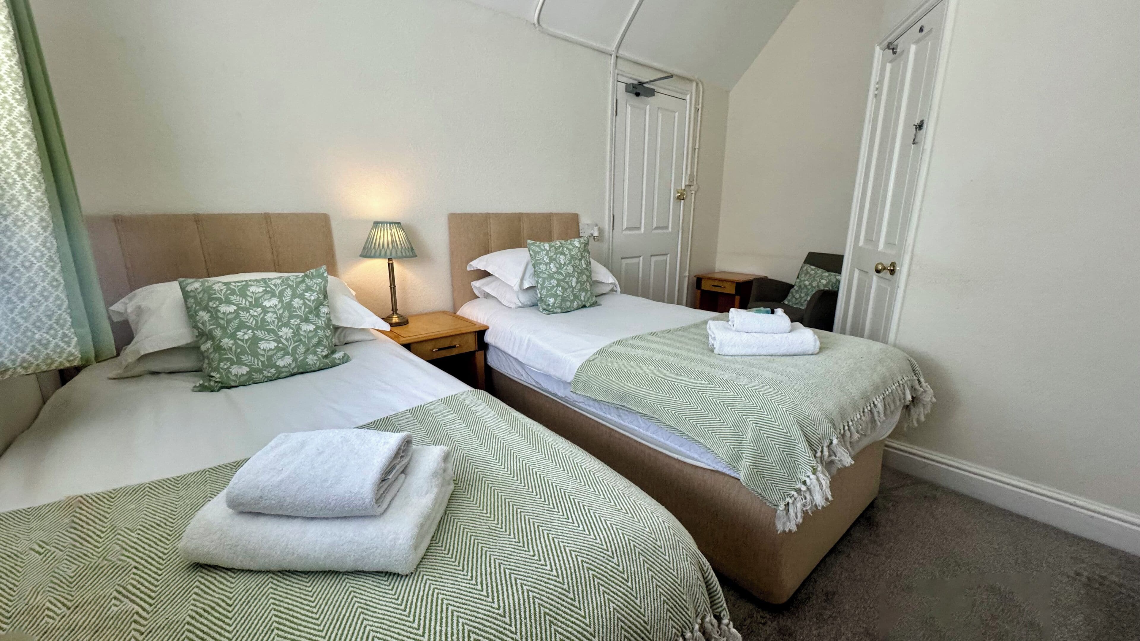 Habitación de hotel con dos camas, cada una con cubrecama y almohadas estampadas en verde, una mesita de noche con lámpara y una puerta con tirador de latón.