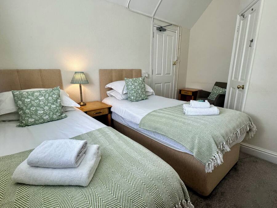 Habitación de hotel con dos camas, cada una con cubrecama y almohadas estampadas en verde, una mesita de noche con lámpara y una puerta con tirador de latón.