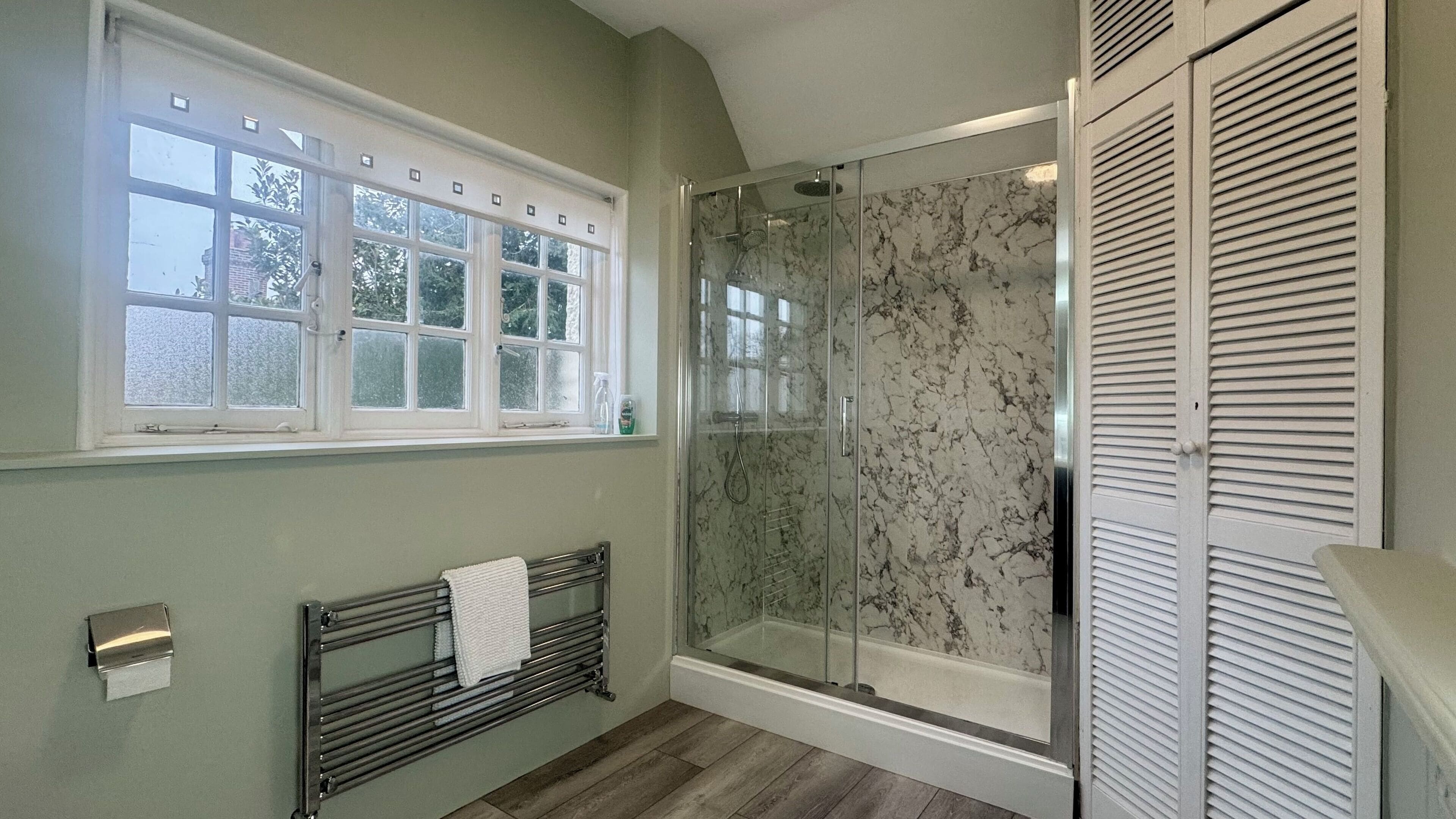 Un baño con ducha envuelta en vidrio, un radiador, una ventana con vidrio esmerilado y un perchero para toallas.