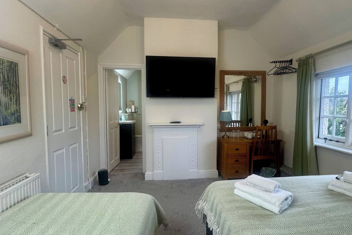 A hotel room with two beds, a TV mounted on the wall, a fireplace, a mirror, a desk with a chair, and a window with curtains.