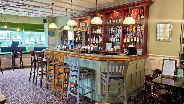 A bar area with a wooden counter, multiple stools, and a variety of bottles and glasses. - The Cricketers Inn (Winchester)