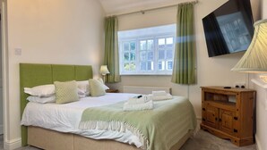 A neatly made bed with a green headboard, white bedding, and a green and white patterned throw. There is a wooden nightstand with a lamp, a wooden cabinet, and a television mounted on the wall. - The Cricketers Inn (Winchester)
