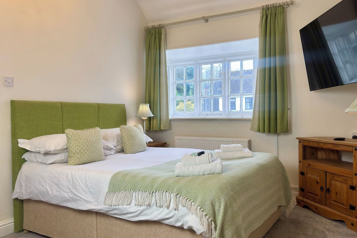 A neatly made bed with a green headboard, white bedding, and a green and white patterned throw. There is a wooden nightstand with a lamp, a wooden cabinet, and a television mounted on the wall.