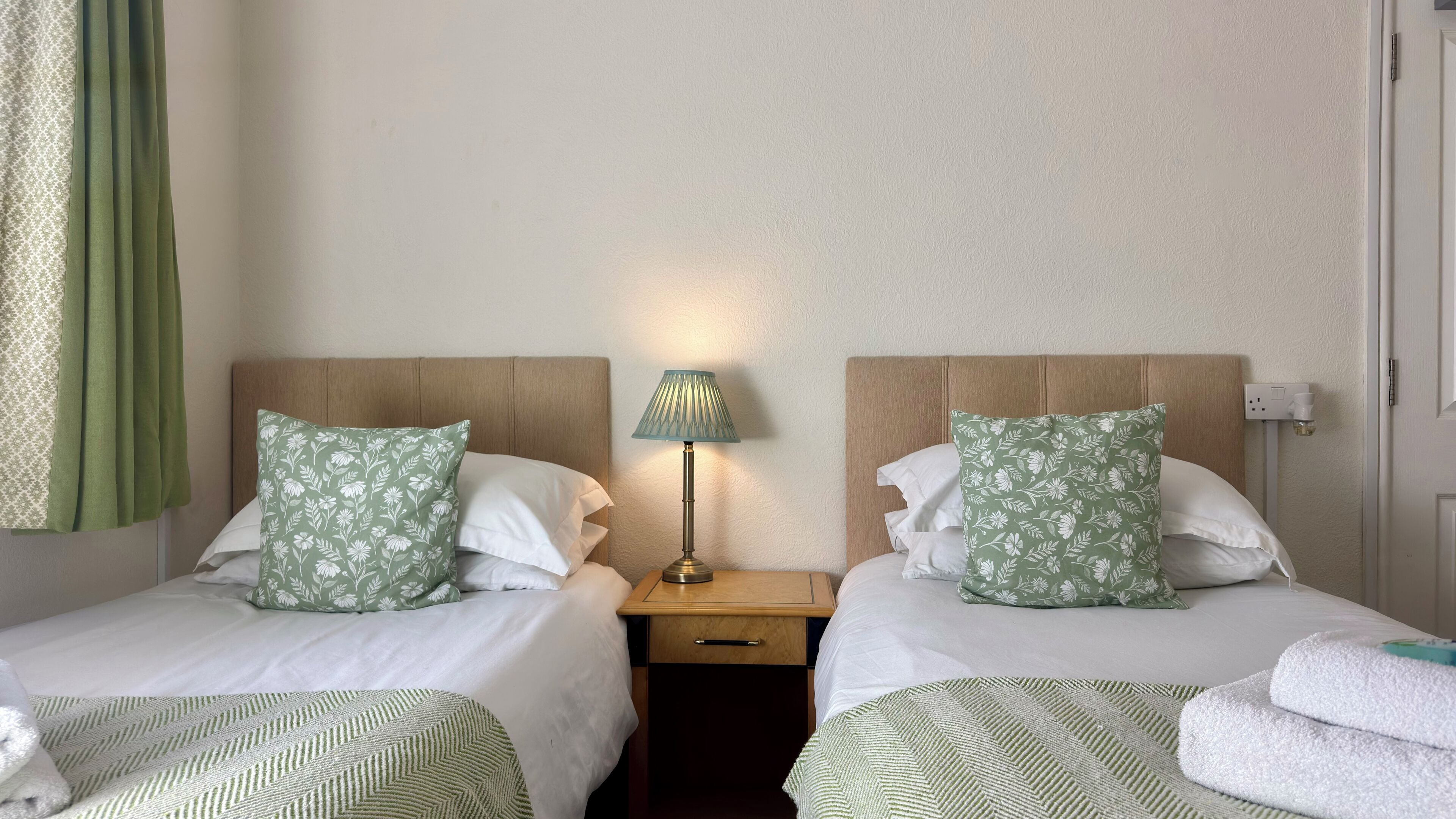 Habitación de hotel con dos camas, cada una con almohadas estampadas de flores verdes, una mesita de noche con lámpara y una manta verde y blanca a rayas.