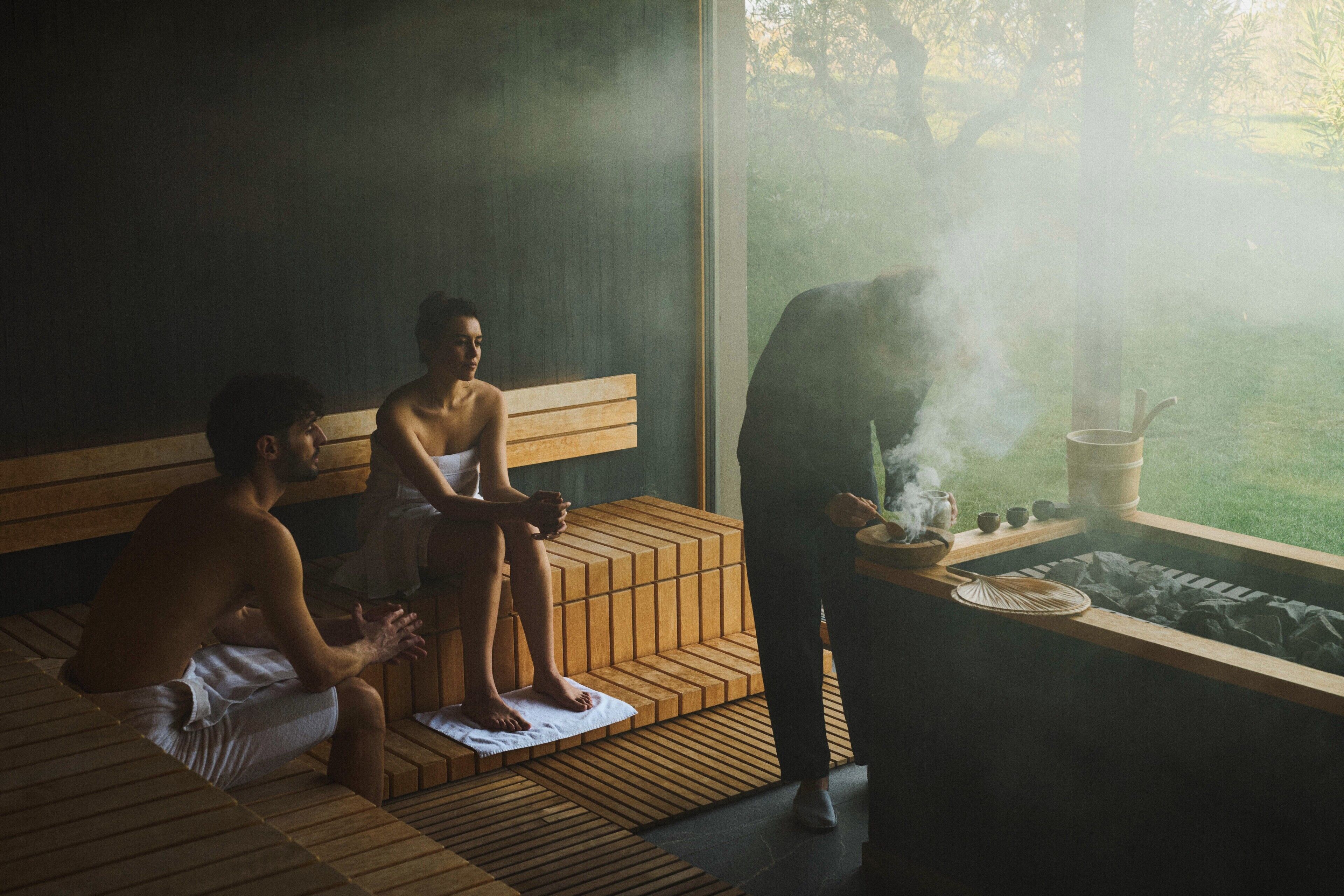 A sauna with wooden benches, a person tending to the steam, and a view of greenery outside.