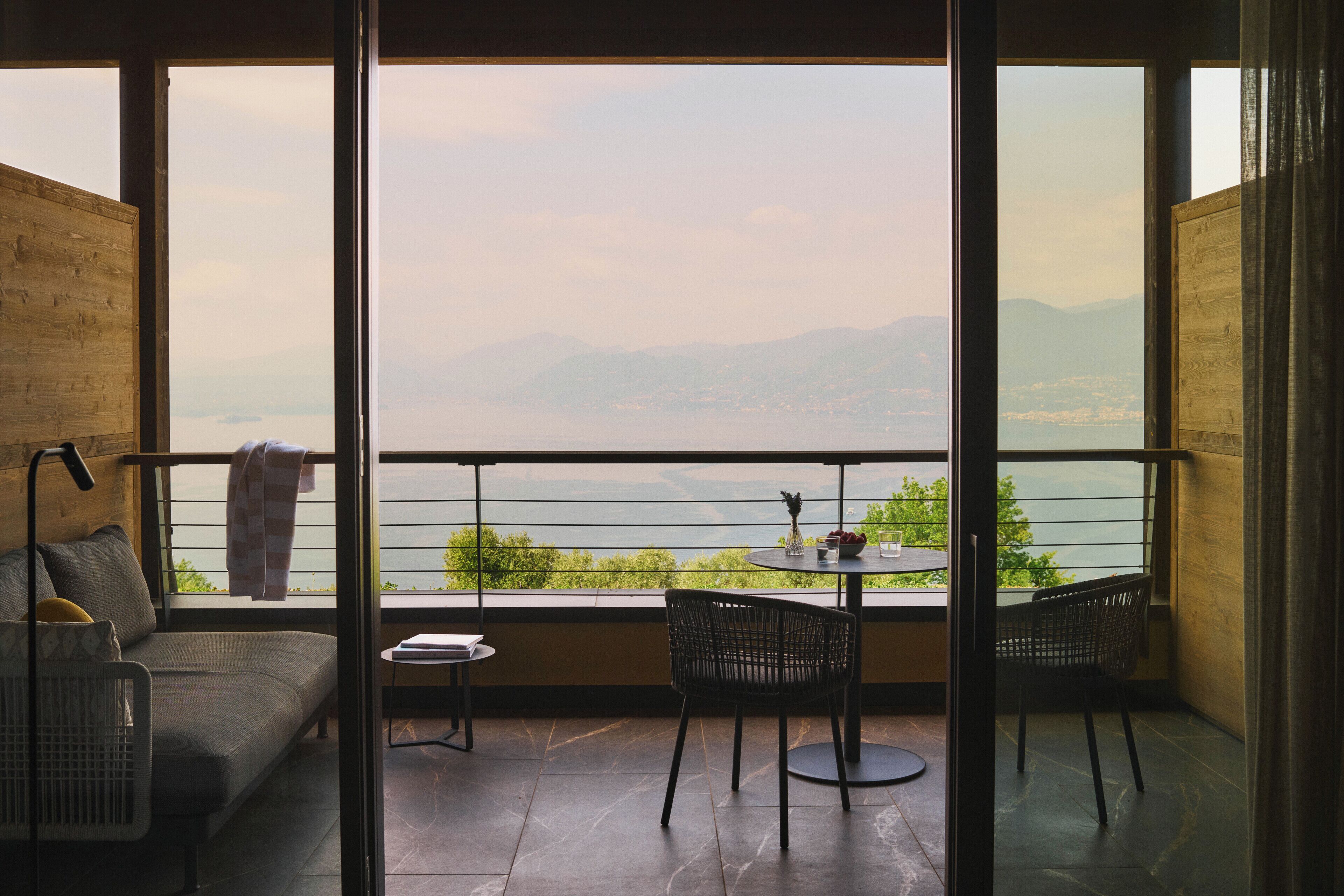 Uma varanda com vista para montanhas e um corpo de água, equipada com um sofá, duas cadeiras e uma mesinha.