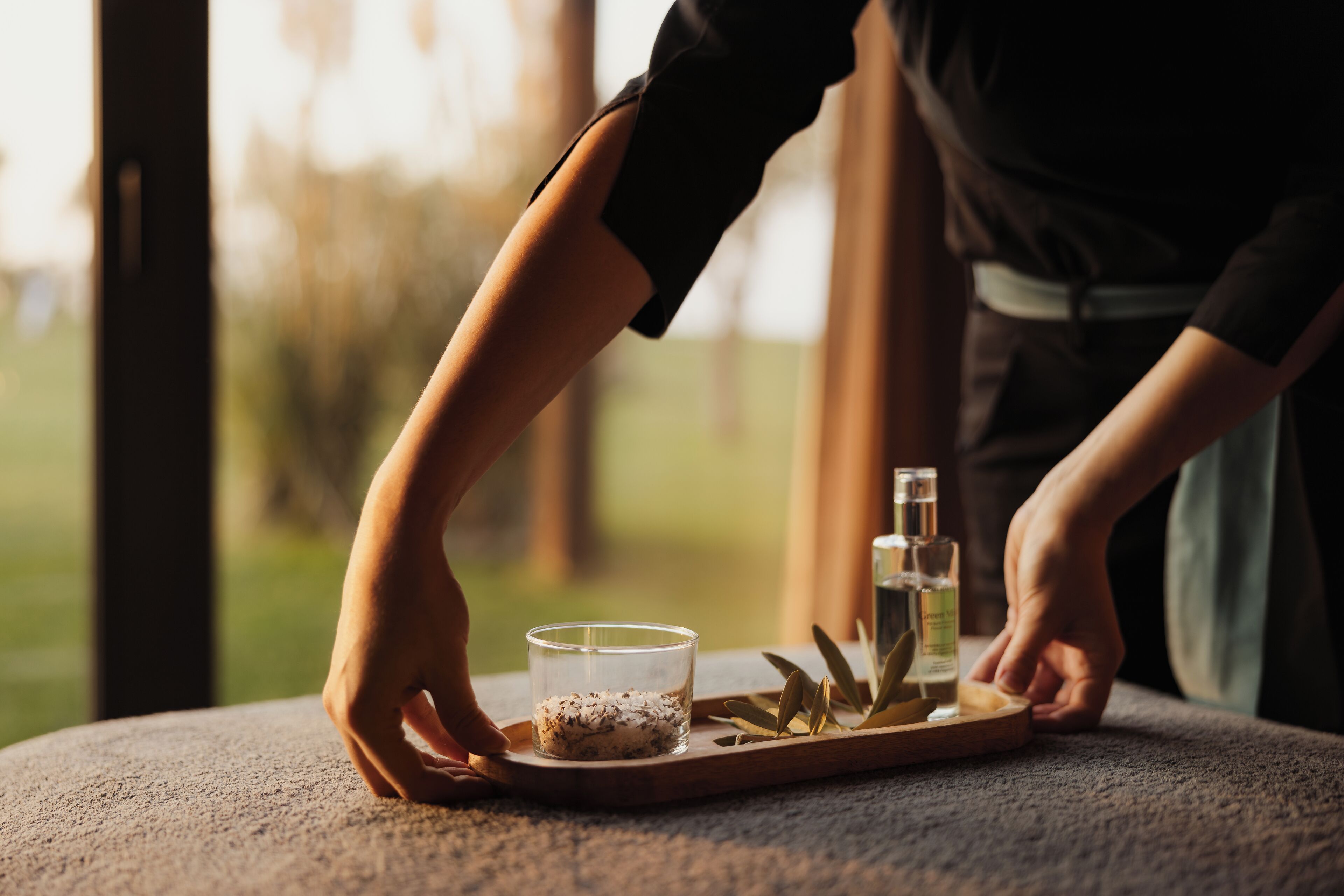 A person placing a tray with a glass, a bottle, and some leaves on a round table.
