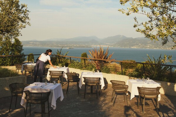 An outdoor dining area with tables and chairs, overlooking a body of water and distant mountains. - Cape of Senses (Torri del Benaco)