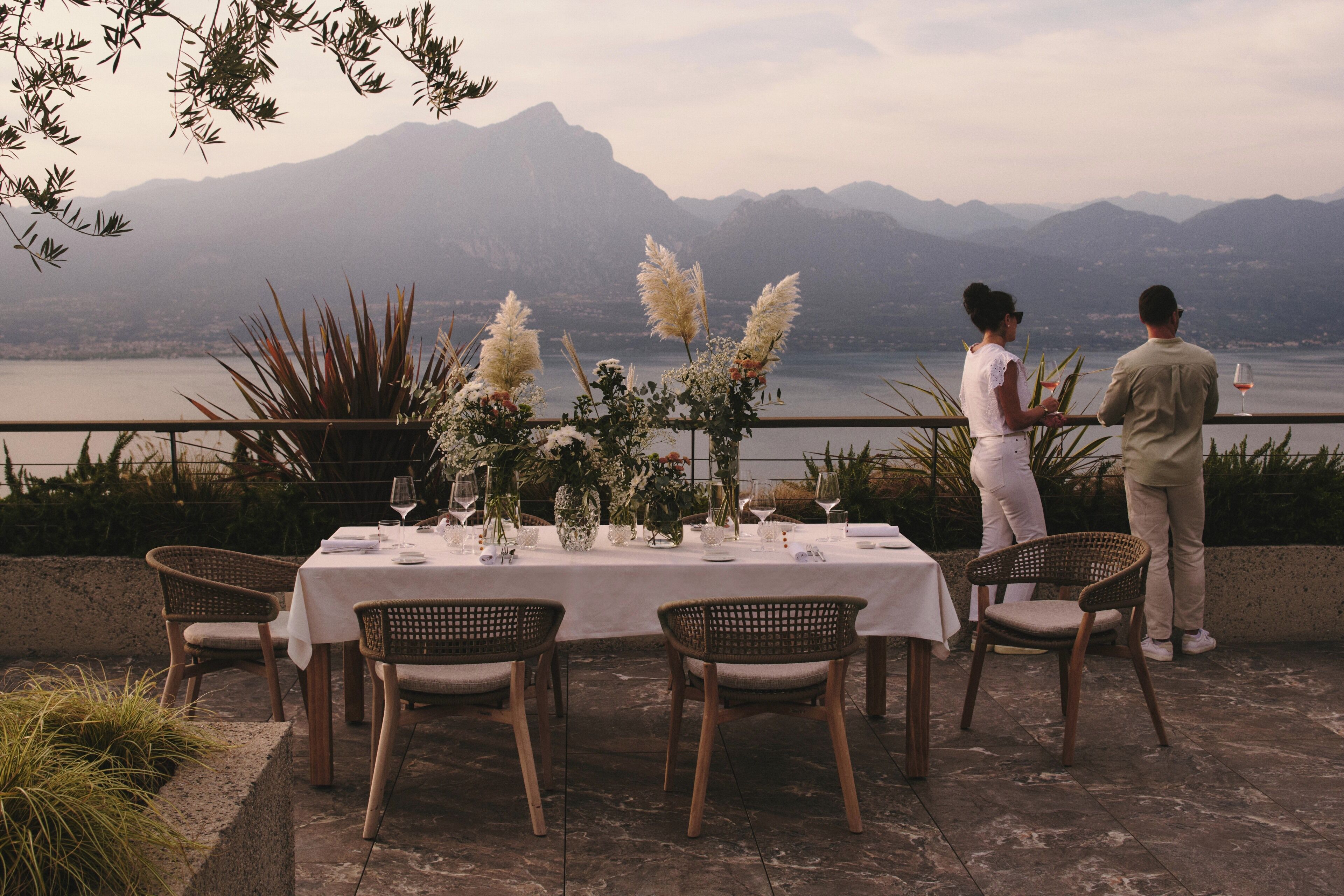 Un espacio de comedor al aire libre con una mesa dispuesta para dos personas, rodeada de sillas de mimbre, con vista a un lago y montañas al fondo.