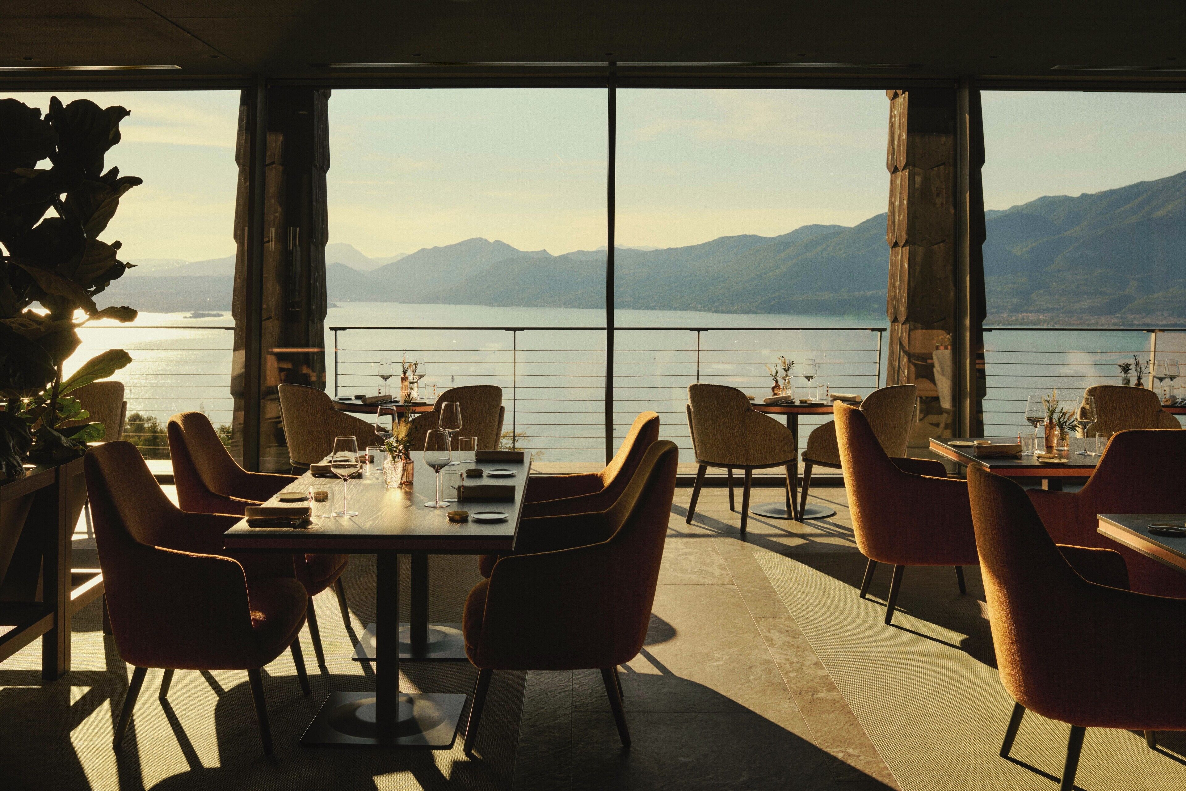 En restaurant med borde og stole, indrettet til spisning, med udsigt til en sø og bjerge i baggrunden.
