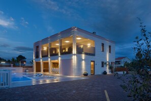 A modern two-story building with a swimming pool, large windows, and a well-lit entrance.