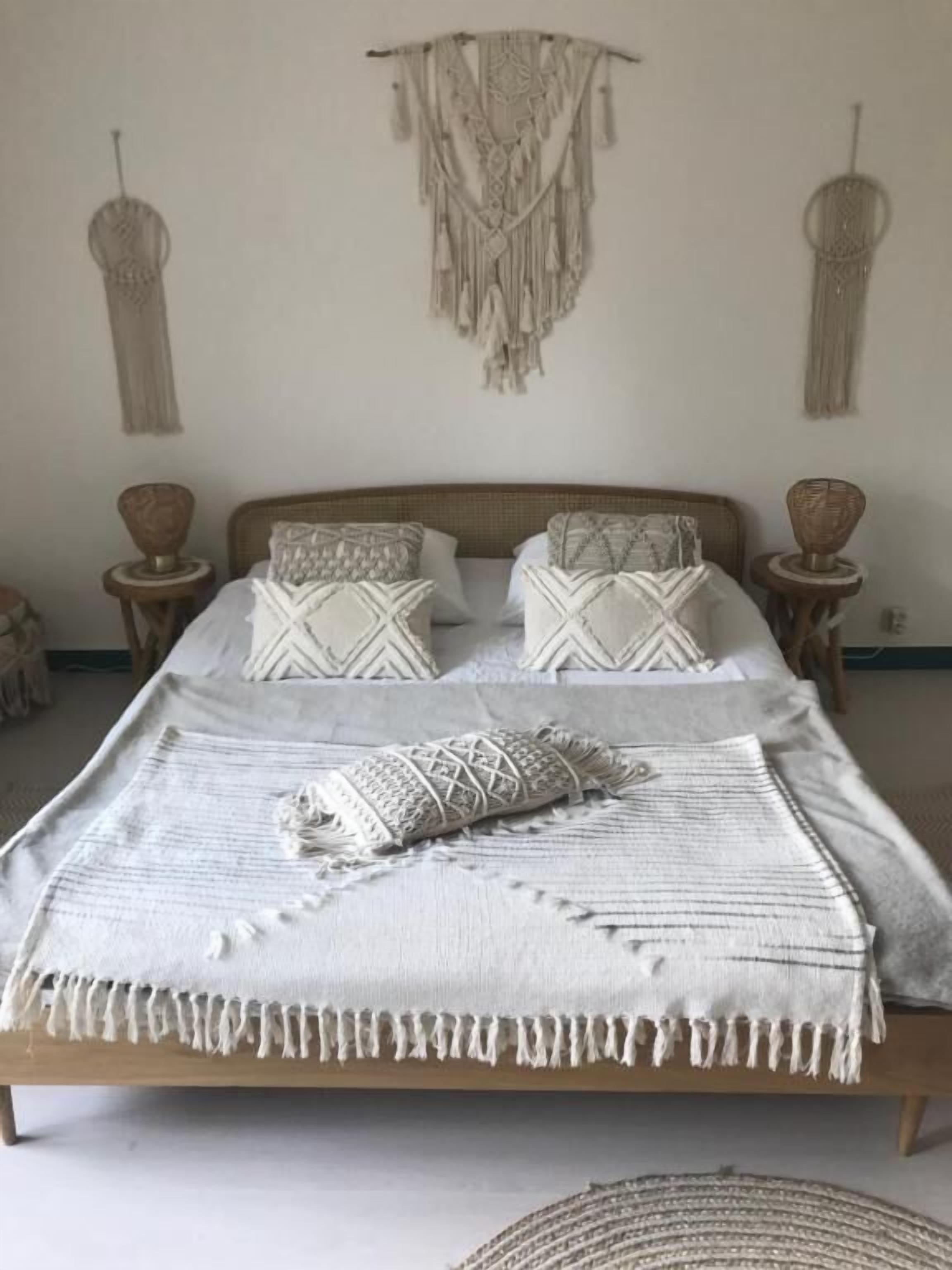 Un lit avec un cadre en bois, une literie blanche et des coussins décoratifs. Au-dessus du lit, on peut voir des décorations murales en macramé.