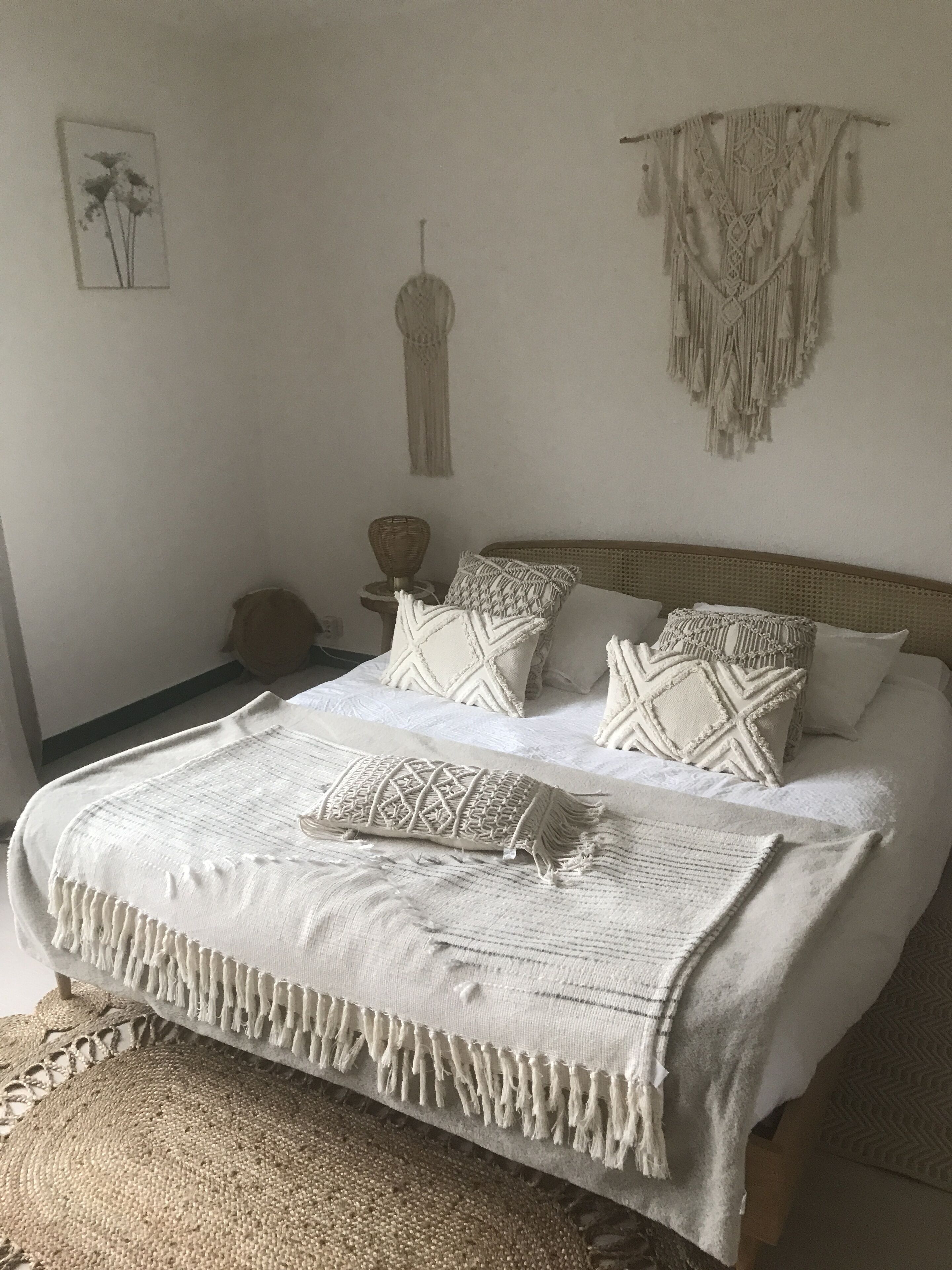 Un lit recouvert d’une courtepointe blanche et agrémenté de plusieurs oreillers, une décoration murale en macramé et un tableau encadré accroché au mur.