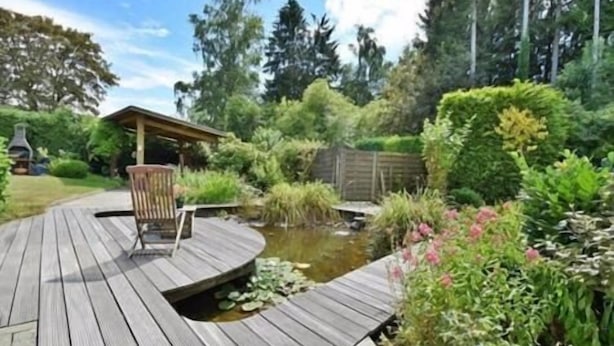 Una terraza de madera con un estanque, rodeada de abundante vegetación y una cerca de madera.