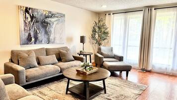 A modern living room with a sofa, armchair, and coffee table, a painting on the wall, and a large window with curtains.