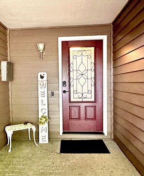 A red door with a decorative glass panel, a welcome sign, a small bench, and a wall-mounted light fixture.