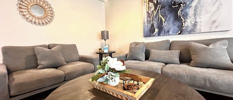 Un salon avec deux canapés gris, une table basse ronde agrémentée d’un plateau décoratif, un miroir mural et un grand tableau abstrait.