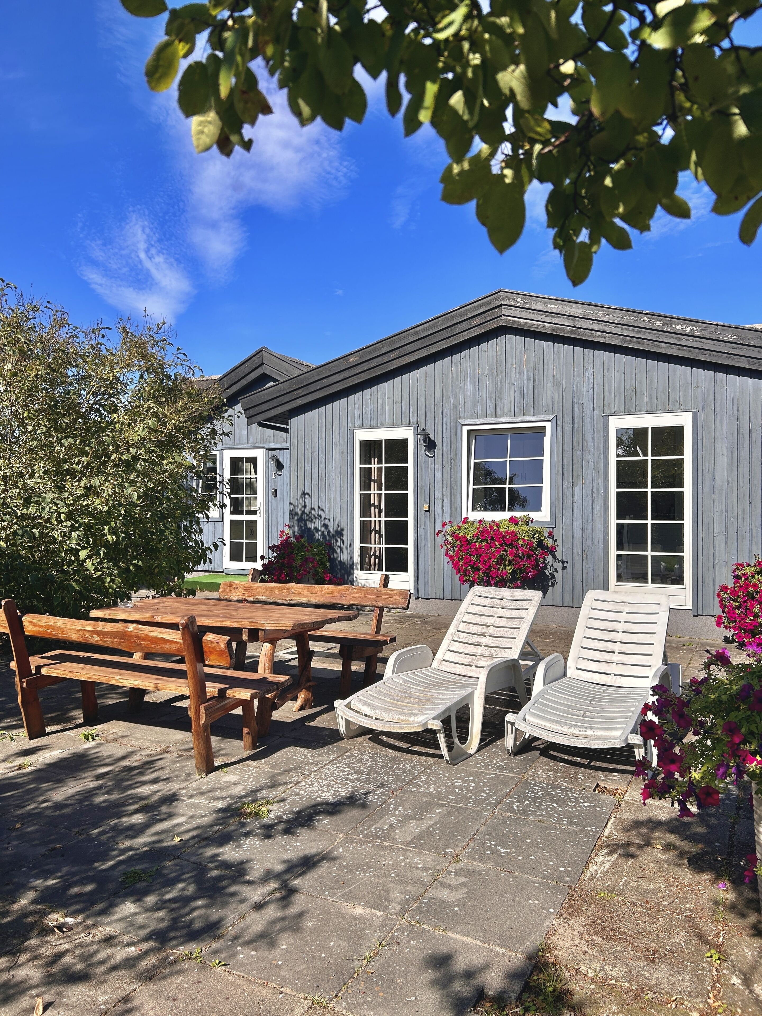 En byggnad med en grå fasad, vita fönster och en uteplats med träbänkar och vita loungestolar.