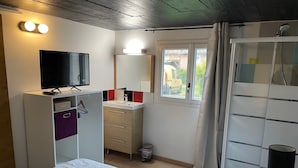 Ein Schlafzimmer mit Bett, Fernseher, Kleiderschrank, Bad mit Dusche und einem Fenster mit Vorhängen.